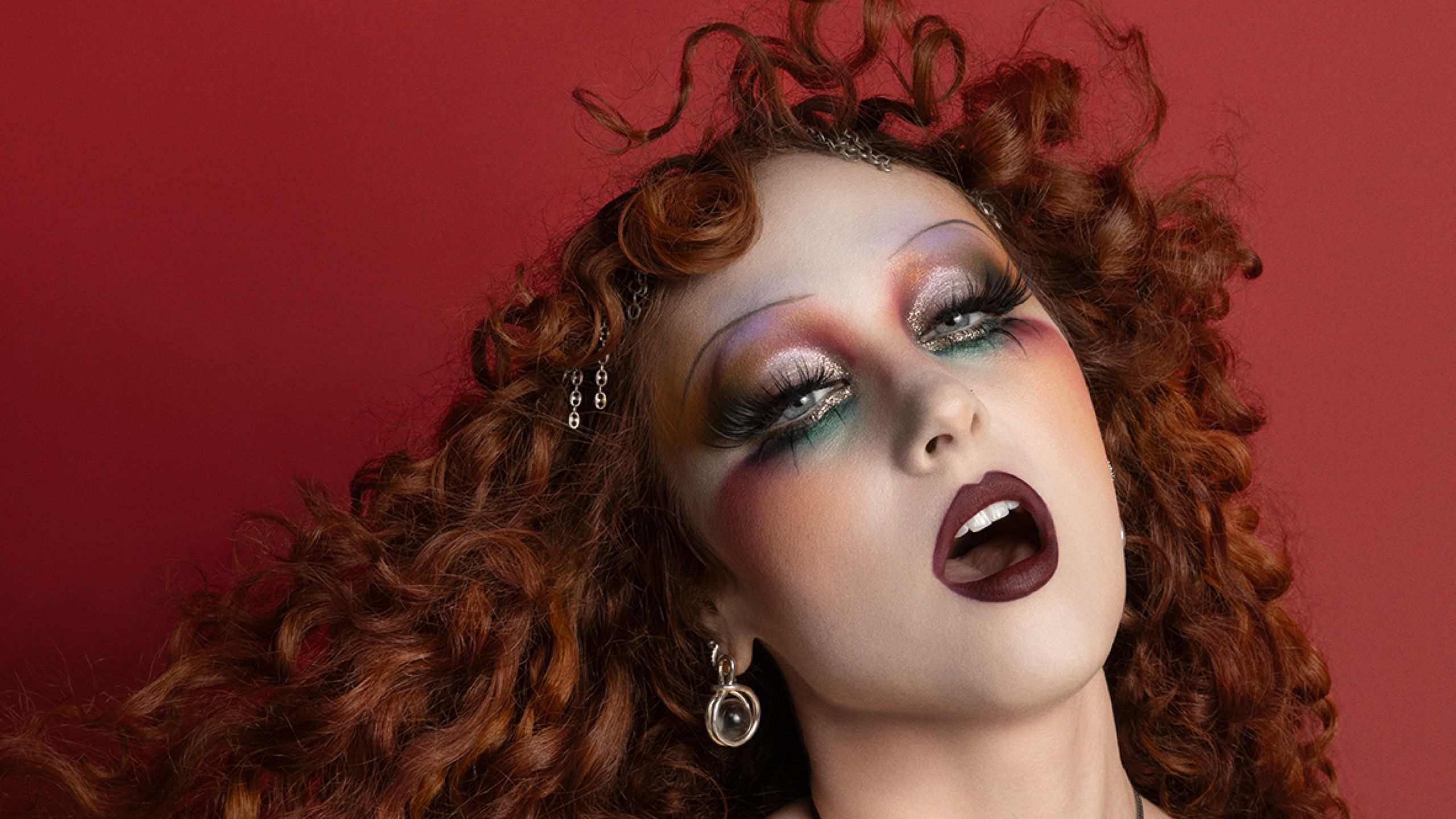 Chappell Roan appears in a MAC Viva Glam ad wearing a flowing costume and holding a flag. Her hair is styled in curls...
